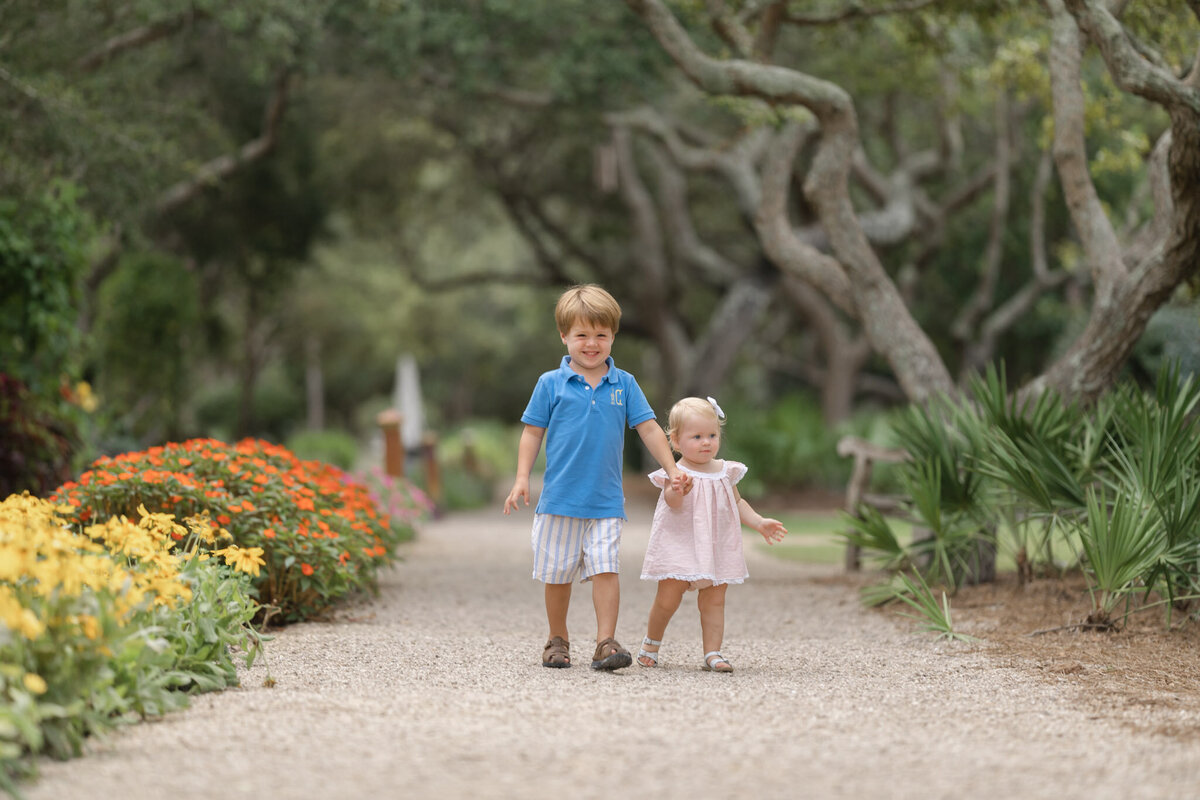 A big brother in a blue polo walks down a flower garden gravel path holding the hand of his toddler sister in a pink dress fancy pants charlotte