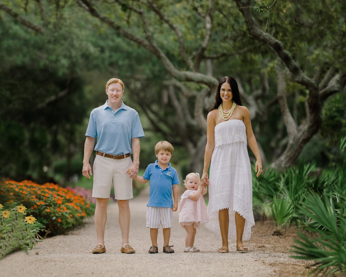 A family of four holds hands while walking down a gravel park path in a flower garden fancy pants charlotte
