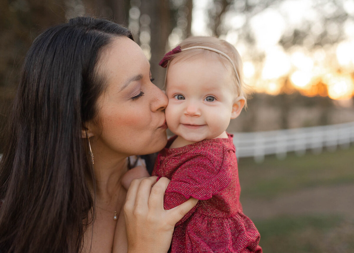 A mother stands in a field at sunset kissing the cheek of her toddler daughter in a red dress polliwogs greensboro
