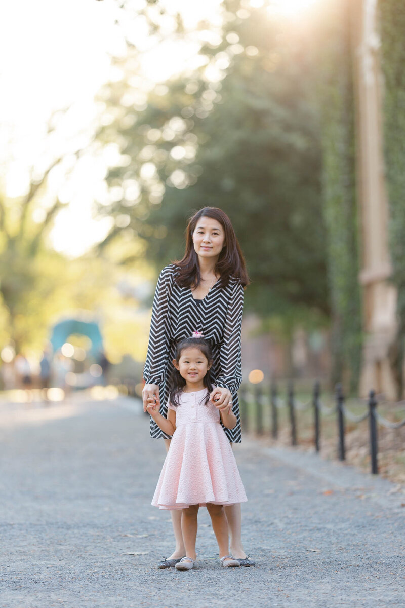 A mother in a stripped dress stands in a park path holding hands with her daughter in front of her in a pink dress things to do in charlotte with kids