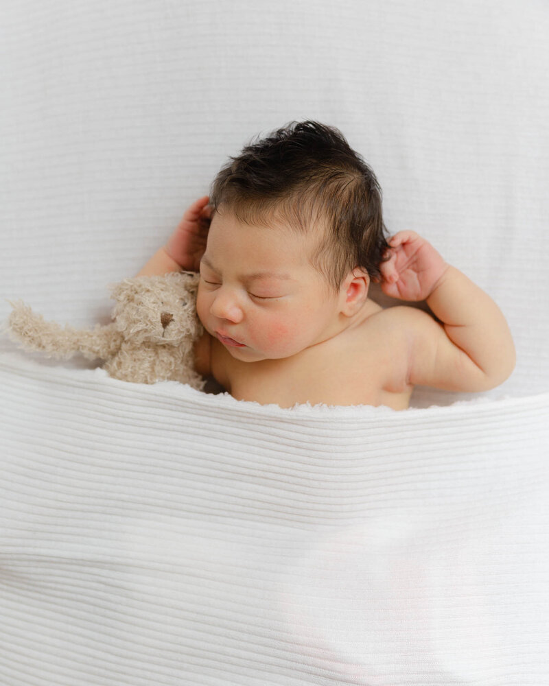 A newborn baby sleeps under a white blanket with a stuffed bear