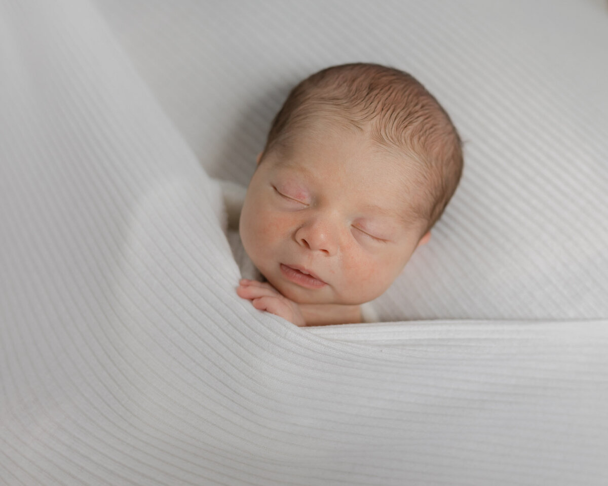 A newborn baby sleeps in a white blanket with a hand sticking out